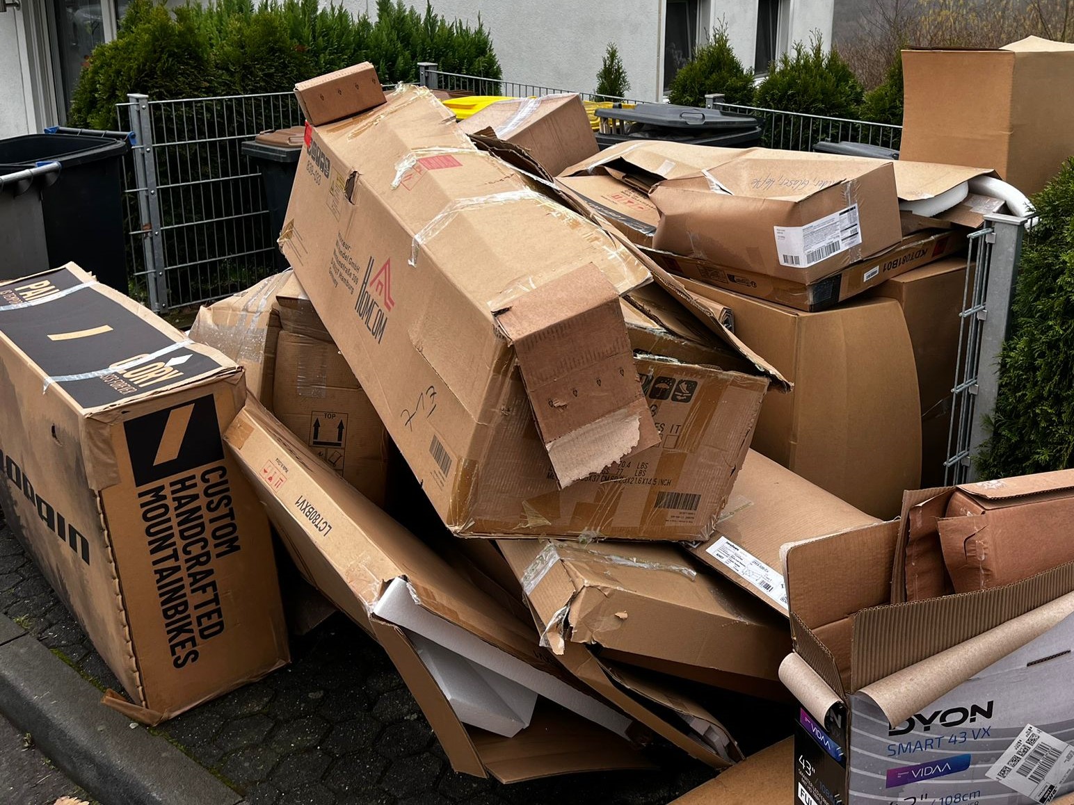 Eine Szene am Straßenrand, wo ein Stapel von Kartons wahllos gestapelt ist. Die Kartons variieren in Größe und Form, einige sind mit Aufklebern und Preisschildern versehen. Im Hintergrund ist eine Mülltonne zu sehen 